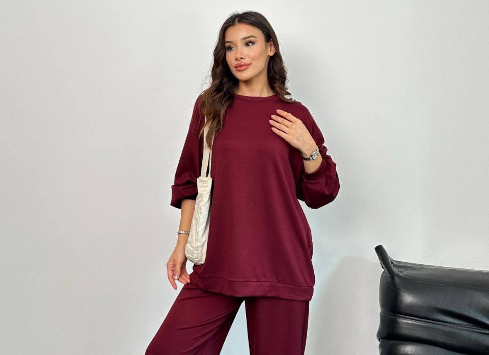 Woman wearing a burgundy outfit standing next to a black leather chair against a white wall.